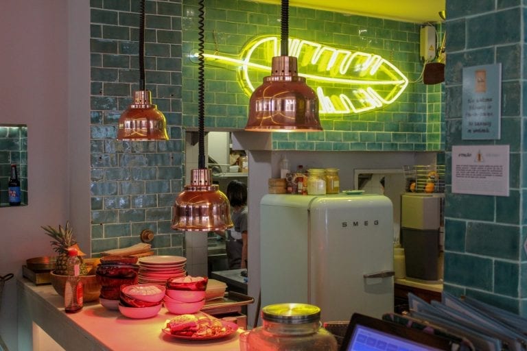 Imagem mostra o balcão e parte da cozinha de uma lanchonete, com luzes acesas, geladeira e outros utensílios de cozinha.