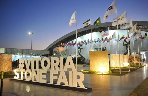 Foto de anos passados do letreiro da Vitória Stone Fair, com pavilhão de exposição ao fundo e bandeiras